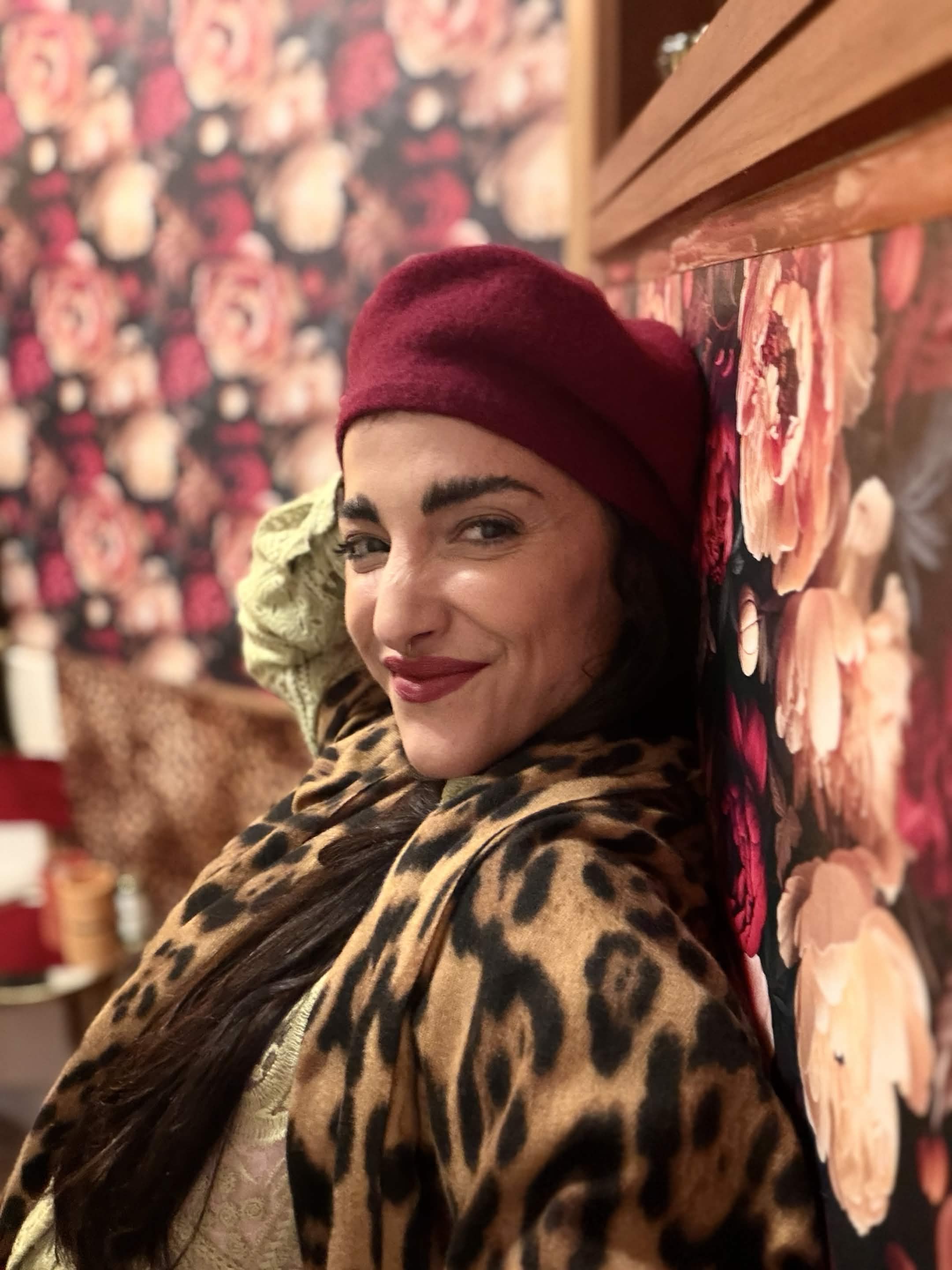 Aline sentada no sofá com boina vinho, estampa animal print e parede floral ao fundo.